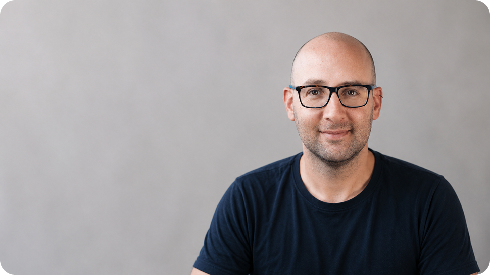 Portrait of a bald man with glasses wearing a navy T-shirt against a gray backdrop.